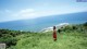 A woman in a red dress standing on a grassy hill overlooking the ocean.